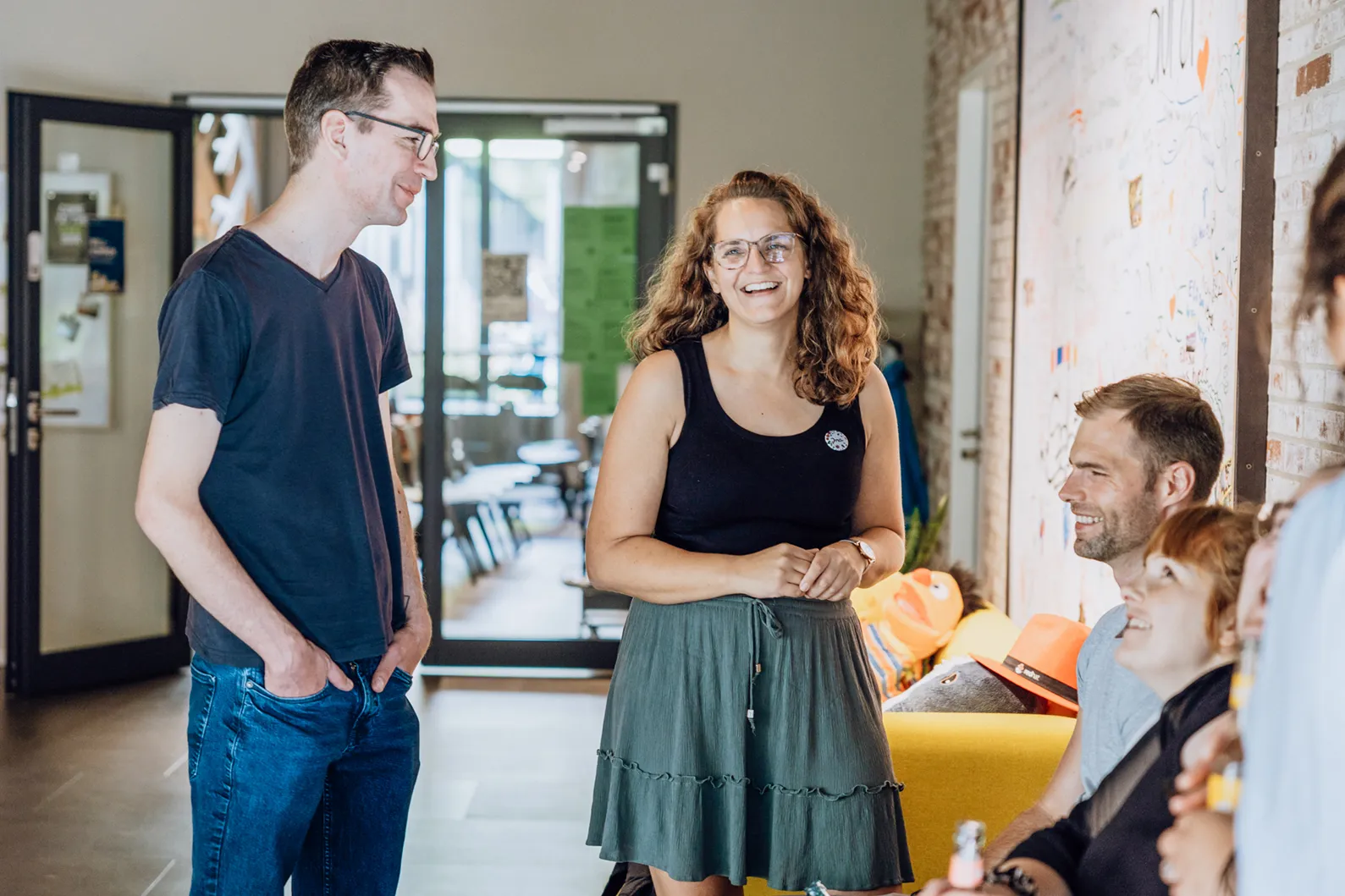 A group of people smiling and chatting in a casual office setting, with a colorful wall in the background.