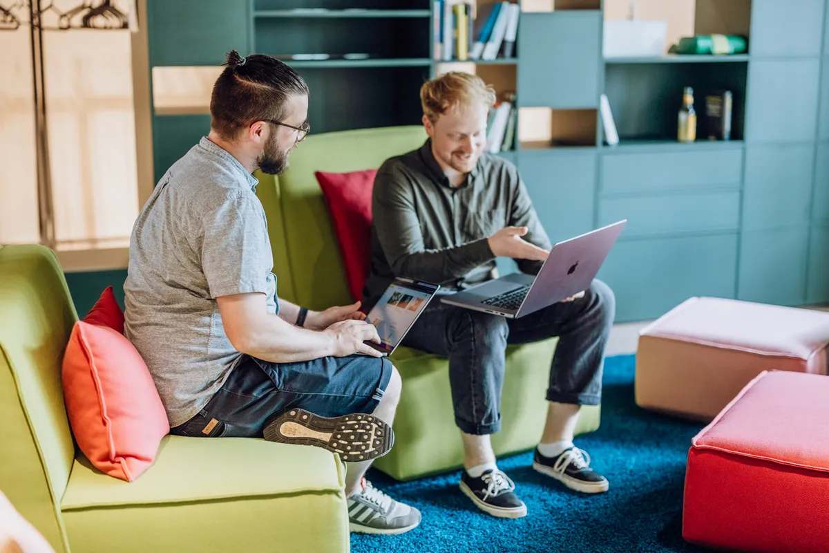 Two men sitting on colorful couches using laptops in a modern office setting.
