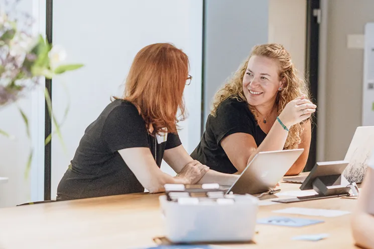 Two women sitting at a table with laptops, engaged in a friendly conversation in a bright office setting.